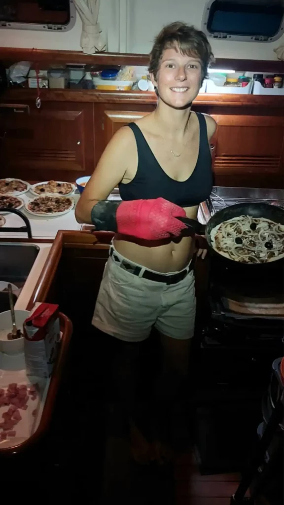 Sophie du Mama Beluga est dans la cabine principale du bateau Mama Beluga est tient dans sa main une poêle avec une pizza. Sur une table en arrière plan, nous pouvons également voir plusieurs pizzas cuite au Levain